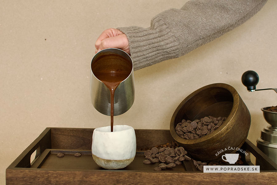 Ruka v pletenom svetri nalieva hustú horúcu čokoládu z kovovej nádoby do bieleho keramického hrnčeka, okolo sú rozliate čokoládové kúsky. Príprava lahodnej horúcej čokolády s kávou