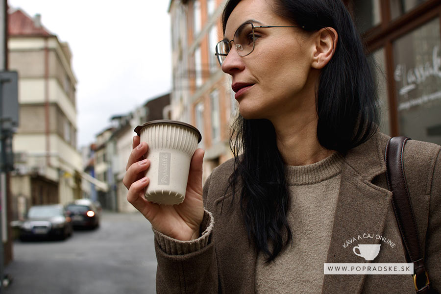 Žena v hnedom kabáte a okuliaroch drží ekologický pohár s viečkom na ulici. Ekologický pohár s horúcou čokoládou do sychravého dňa
